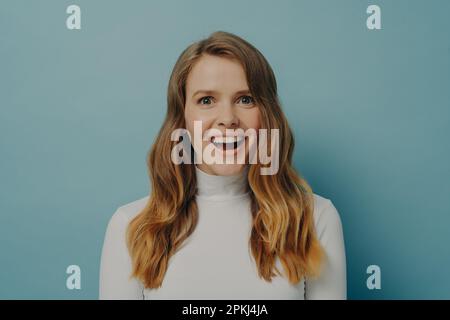 Immagine ravvicinata di bella giovane donna felice in abiti casual con capelli ondulati marrone chiaro guardando la macchina fotografica con il sorriso di fascio ridendo su divertente jo Foto Stock