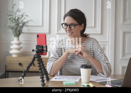 Giovane donna d'affari spagnola sorridente in occhiali seduti davanti allo smartphone sul treppiede sul posto di lavoro, influencer positivo femmina che registra video per b Foto Stock