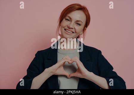 Scatto isolato da studio di giovane donna sorridente e rossa che fa forma al cuore con entrambe le mani e sorridendo dolcemente alla macchina fotografica, dimostrando amore e cura Foto Stock