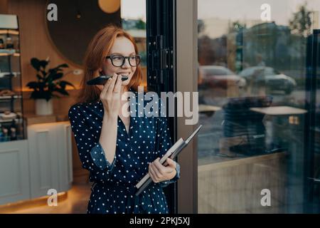 Sorridente e penosa giovane donna rossa effettua chiamate vocali tramite smartphone guarda via stand alla porta del bar utilizza moderne tecnologie per la comunicazione Foto Stock