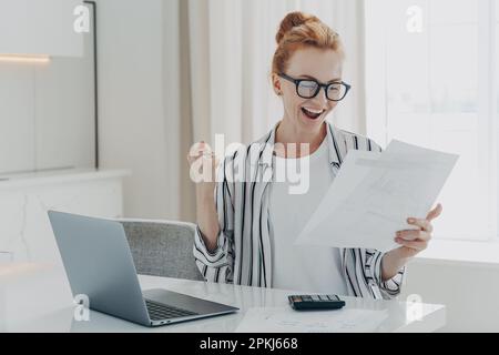 Emotivo felice giovane donna zenzero in eyewear tenendo carta con l'ultima notifica bancaria pagamento ipotecario, sollevando la mano con pugno clenched, lettura Foto Stock