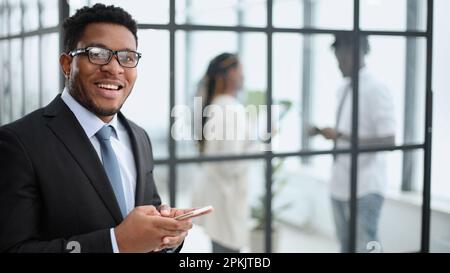 Uomo d'affari afro-americano in un elegante vestito nero guarda la fotocamera sullo sfondo dei colleghi Foto Stock