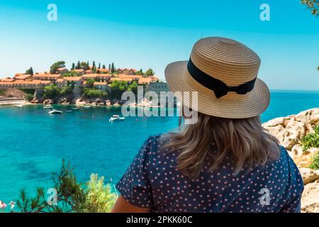 Montenegro. Mare Adriatico. Isola e spiaggia di Sveti Stefan. Tempo soleggiato. Un luogo turistico molto popolare. Donna in un cappello di paglia ammirando una bella vista Foto Stock