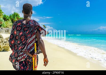 Uomo Maasai con abiti tradizionali e bastone ammirando il mare cristallino in piedi su una spiaggia, Zanzibar, Tanzania, Africa orientale, Africa Foto Stock