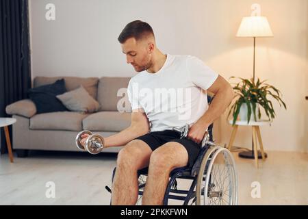 Fare esercizi utilizzando manubri. L'uomo disabile in sedia a rotelle è a casa. Foto Stock