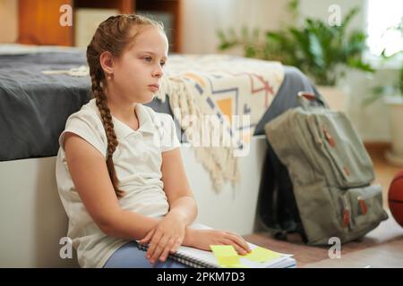Scolaretta stanca dopo aver fatto i compiti seduti sul pavimento accanto al letto Foto Stock