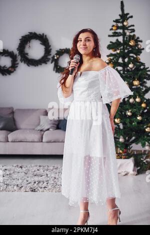 Donna in vestito bianco e con microfono in mano sta cantando nella stanza domestica decorata di natale. Foto Stock