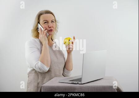 donna che parla al telefono è turbata e dispiaciuta nelle sue mani ha la dieta di mela corretta ricette di menu nutrizionale di fronte al portatile Foto Stock