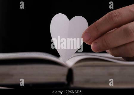 Libro con pagine aperte e carta a forma di cuore. Donna mano che tiene il cuore a forma di carta su libro aperto sul tavolo. amore per la lettura concetto Foto Stock