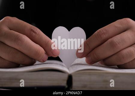 Libro con pagine aperte e carta a forma di cuore. Donna mano che tiene il cuore a forma di carta su libro aperto sul tavolo. amore per la lettura concetto Foto Stock