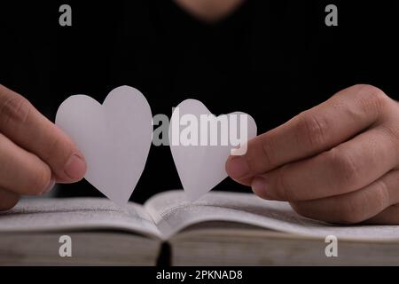 Libro con pagine aperte e carta a forma di cuore. Donna mano che tiene il cuore a forma di carta su libro aperto sul tavolo. amore per la lettura concetto Foto Stock