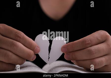 Mano femminile che tiene un cuore di carta bianca mezzo-strappato sopra un libro aperto sul tavolo. Cuore di carta bianca strappato a metà in mano Foto Stock