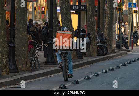 Consegna uomo in bicicletta con uno zaino arancione con la schiena girata su una pista ciclabile a Barcellona lungo una strada fiancheggiata da alberi e persone a piedi Foto Stock