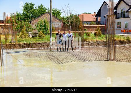 Zrenjanin, Vojvodina, Serbia - 21 luglio 2022: Tre operai di costruzione che abbracciano sorridono e si posano accanto al tubo flessibile della pompa come gettando cemento fresco Foto Stock