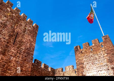 Le due imponenti torri principali di Castelo de Silves, un'antica fortezza moresca con massicce mura in pietra, mostrano con orgoglio una bandiera portoghese che sventola Foto Stock