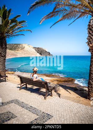 Una giovane donna irriconoscibile che indossa un cappello mentre siede su una panchina tra palme, godendosi la vista sulla spiaggia Praia da Luz con Rocha Negra rock. Foto Stock
