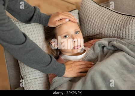 Preoccupatevi donna millenaria che tocca la fronte della triste ragazza teen europea, che soffre di malattia, si trova sul divano Foto Stock