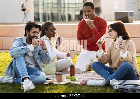 Sorridenti studenti internazionali moderni studiano insieme, si riposano dalla lezione, si siedono sull'erba, mangiano cibo Foto Stock