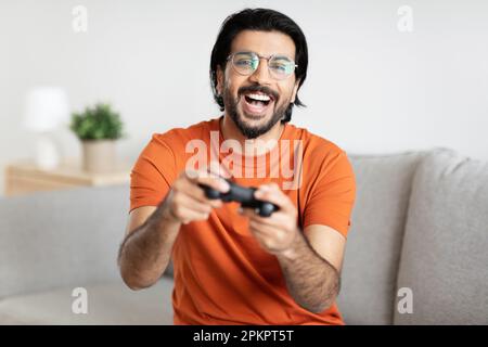 Felice uomo giocatore arabo millenario in occhiali con barba si siede sul divano con joystick, gioca online da solo Foto Stock