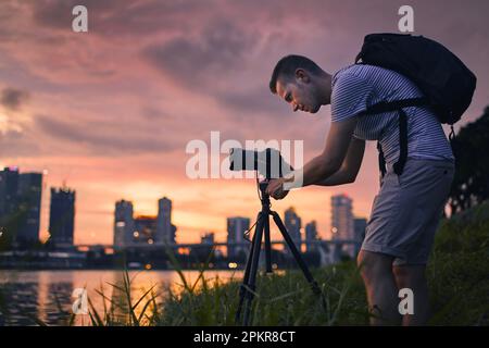 Fotografo concentrato con fotocamera su cavalletto durante lo scatto di foto dello skyline urbano. Città moderna durante il bellissimo tramonto, Singapore. Foto Stock