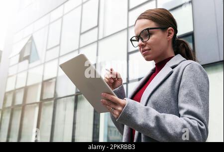 Una donna d'affari che indossa gli occhiali si trova accanto al business center e lavora su un tablet digitale. Foto Stock