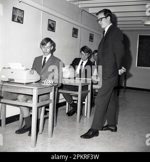 1960s, studenti di college maschi, storici e ben vestiti, seduti alle scrivanie utilizzando macchine da scrivere elettriche, sotto la supervisione di un giovane insegnante o supervisore di sesso maschile adulto in una classe di dattilografia, Middletown, USA. Il modello di macchine da scrivere utilizzato è la macchina da scrivere IBM Selectric, introdotta per la prima volta sul mercato nel 1961. Foto Stock