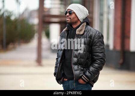 Ritratto di un giovane bell'uomo nero, in piedi sul paesaggio urbano, guardando lontano, indossando giacca in pelle, cappello lavorato a maglia e occhiali da sole. Moda Street style Foto Stock