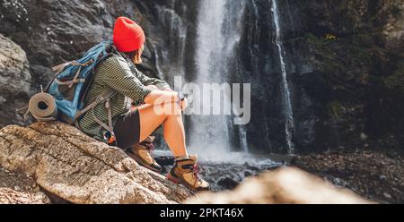 Donna con uno zaino in un cappello rosso vestito con abiti da trekking attivi seduti vicino alla cascata del fiume di montagna e godersi gli spruzzi di natura Foto Stock