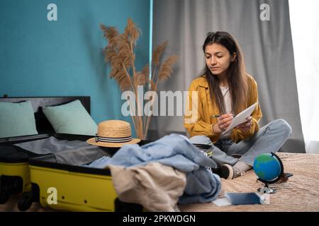 Concetto di vacanza di viaggio. Donna che fa la lista di controllo delle cose da imballare per il viaggio, la scrittura della nota di presa della carta, la valigia di imballaggio alla vacanza che si siede sul Foto Stock