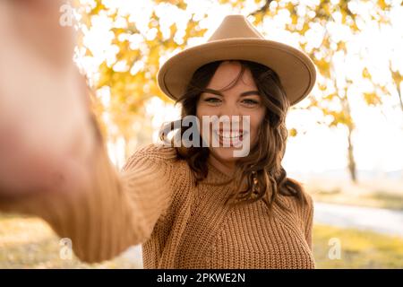 Donna sorridente in maglione alla moda divertirsi nel parco autunnale. Adorabile ragazza che prende selfie su sfondo sfocato autunno natura. Spazio di copia Foto Stock