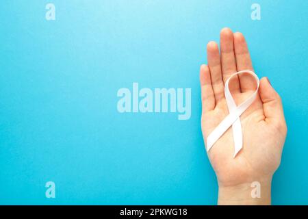 Mano che tiene il nastro bianco su sfondo blu. Concetto di consapevolezza del cancro polmonare. Vista dall'alto Foto Stock