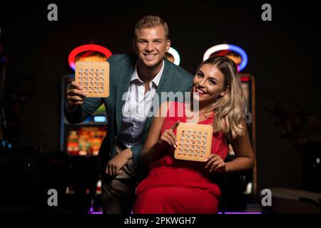 Ritratto di una giovane coppia bella e ben vestita giocando nel Casinò e celebrare Holding Bingo Wood Game Card Foto Stock