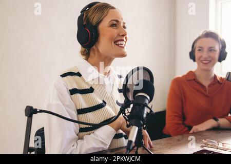 Felice ospite del podcast femminile sorridente mentre presenta un ospite sul suo programma. Due donne che registrano una trasmissione radiofonica in uno studio domestico. Influencer femminili cre Foto Stock