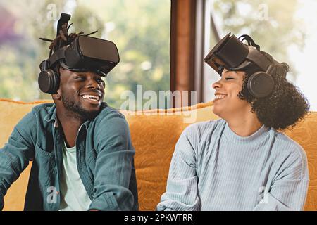 Due giovani amici, un afro-americano e una brasiliana, ridono insieme indossando i visore VR, seduti su un divano con una finestra e verde Foto Stock