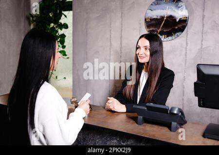 Un giovane e cordiale portiere si trova dietro un banco del ricevimento e fornisce informazioni sulle camere agli ospiti che effettuano il check in in un hotel. Foto Stock