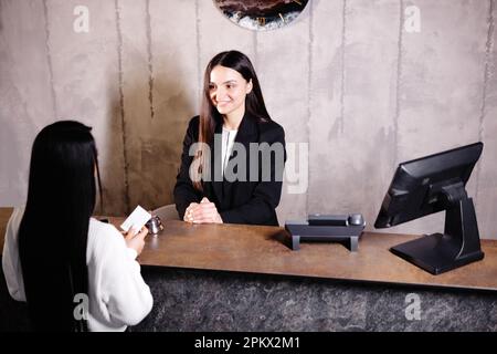 Un giovane e cordiale portiere si trova dietro un banco del ricevimento e fornisce informazioni sulle camere agli ospiti che effettuano il check in in un hotel. Foto Stock