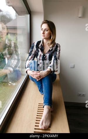 giovane donna in abiti di casa si siede da sola sul davanzale in pensiero Foto Stock