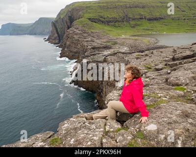 Un turista in giacca rossa si trova su una costa rocciosa sopra la cascata di Bøsdalafossur, che scorre dal lago Sørvágsvatn / Leitisvatn nell'Atlantico Oc Foto Stock