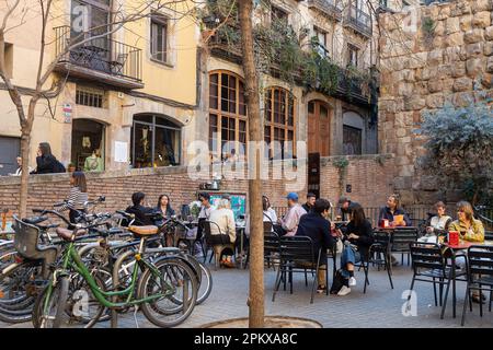 Barcellona, Spagna - 13 marzo 2023: Persone sedute fuori al bar in una piccola piazza. Edifici tradizionali e albero decorativo Foto Stock