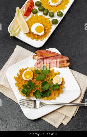 Pancetta, waffle, uova fritte, verdure, olive e una forchetta su due piatti bianchi. Tovagliolo beige sotto il piatto. Giacitura piatta. Sfondo nero. Foto Stock
