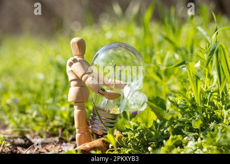 Un piccolo uomo di legno tiene una lampadina incandescente sull'erba nei parchi in Ucraina , elettricità Foto Stock