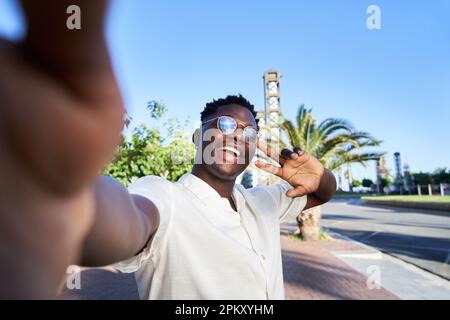 Felice giovane africano che prende un selfie guardando la macchina fotografica e agitando esso, condivisione di social media. Foto Stock