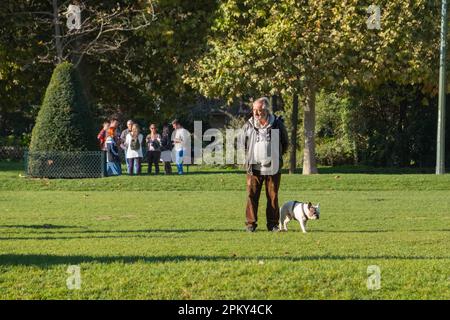 Un uomo di mezza età che cammina con il suo bulldog francese in una giornata di sole in un lussureggiante giardino di Parigi, godendosi la pace all'aria aperta e il legame con il suo animale domestico. Foto Stock