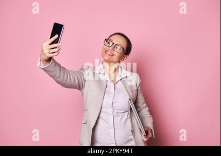 Attraente donna incinta, persona d'affari che indossa abiti casual e occhiali alla moda, che trasporta il laptop e che prende selfie Foto Stock
