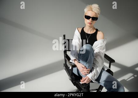 donna bionda elegante in occhiali da sole neri e outfit alla moda seduta su sedia pieghevole su sfondo grigio Foto Stock