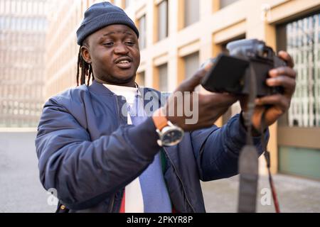 Felice giovane maschio nero in abiti alla moda con trecce in piedi fuori e guardare la fotocamera mentre si scatta foto su una fotocamera professionale Foto Stock