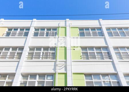 Dal basso di un contemporaneo edificio bianco a più piani con finestre e pannelli verdi sotto il cielo blu senza nuvole nella soleggiata città Foto Stock