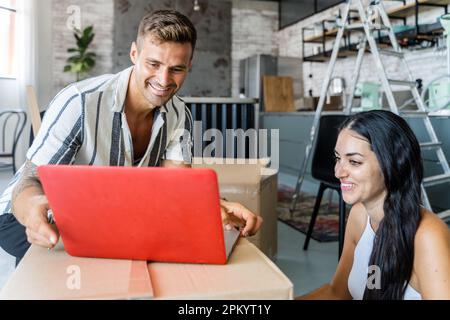 Positivo giovane uomo e donna in abiti casual seduto a scatola di cartone con il computer portatile e guardando lo schermo mentre si sposta in un nuovo appartamento Foto Stock