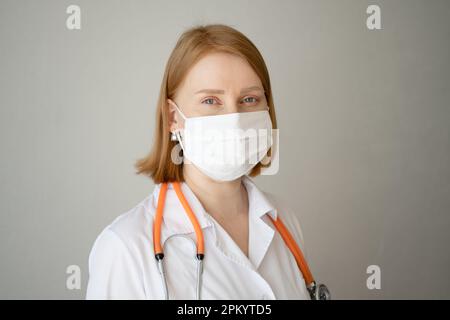 Giovane medico in piedi in mantello medico e maschera protettiva e stetoscopio dietro il collo con guardare la fotocamera contro sfondo grigio Foto Stock