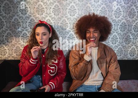 Donna con capelli marroni e ragazzo ispanico positivo seduto vicino alla parete e succhiare lecca mentre si passa il tempo a casa insieme Foto Stock